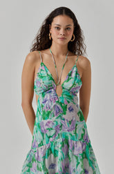 A woman is wearing a sleeveless, flowy dress with thin spaghetti straps. The dress features a vibrant, abstract floral pattern in shades of green, purple, lavender, and white. It has a keyhole cutout at the center of the chest, opening at the bust, and a loose, tiered skirt. She accessorizes with large gold hoop earrings and a long, thin necklace with a circular pendant on the chest.